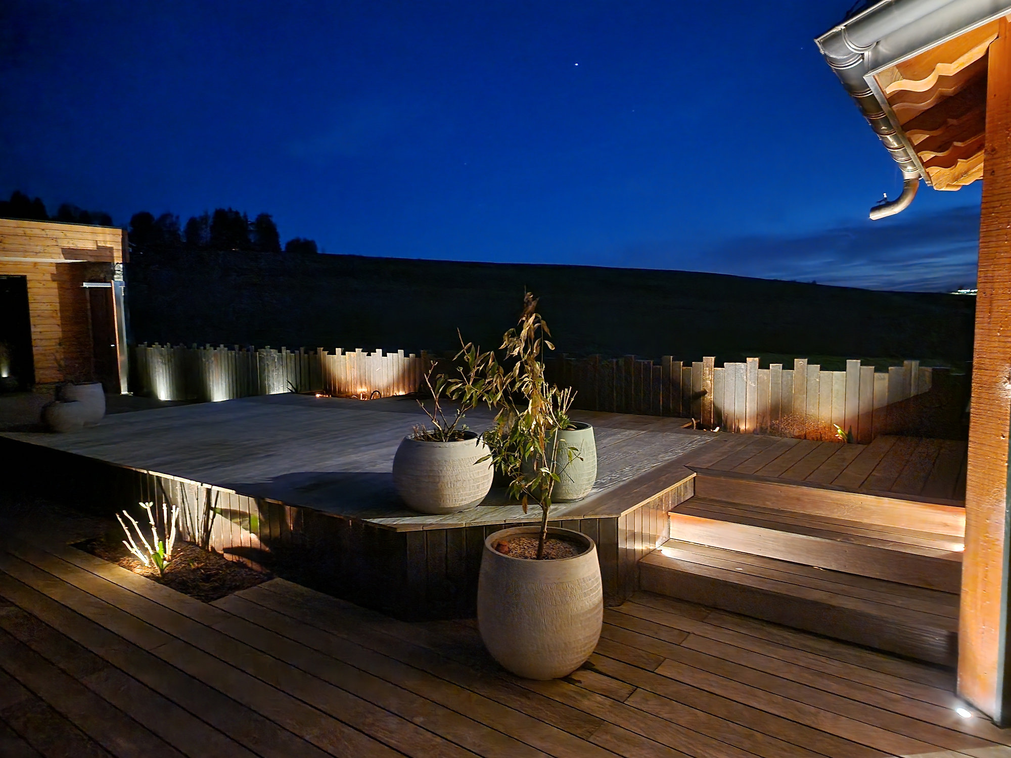Holzterrasse bei Nacht mit beleuchteten Stufen, Pflanzgefässen und angestrahlter Holzpalisade